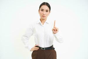 Confident young female manager, asian woman shows number one finger, explains smth, stands over white background photo