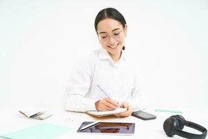 Young asian office woman, sitting at workplace desk, making notes, smiling and looking professional, white background photo