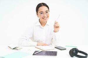 Young saleswoman, asian businesswoman sits in office, works with documents, has digital tablet and headphones on table, white background photo