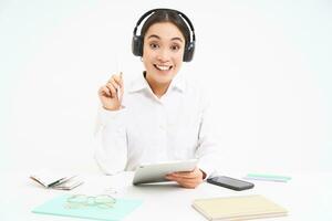 Portrait of professional woman working, listening podcast or course in headphones, holding digital tablet, sitting over white background photo