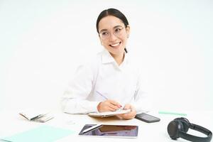 Young asian office woman, sitting at workplace desk, making notes, smiling and looking professional, white background photo