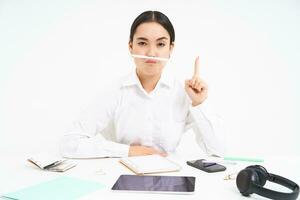 negocio y oficina trabajar. joven mujer sentado a lugar de trabajo, engañando alrededor, sostiene bolígrafo con labios y muestra gracioso caras, blanco antecedentes foto