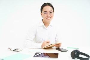 Image of young tutor, asian woman teacher, sits in office, writing, working on report at workplace, wears glasses, white background photo