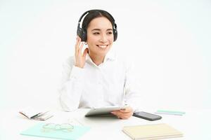 People and workplace. Smiling asian woman sits in office, student listening music in headphones and holding digital tablet, white background photo