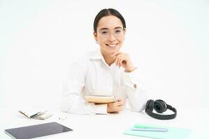 Image of young adult, woman at workplace, sits at workplace in office with documents, headphones and tablet, white background photo