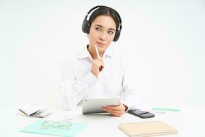 Portrait of professional woman working, listening podcast or course in headphones, holding digital tablet, sitting over white background photo