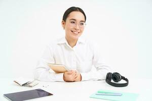 Image of young adult, woman at workplace, sits at workplace in office with documents, headphones and tablet, white background photo