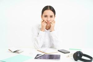 Sad and grumpy asian woman at workplace, sits with digital tablet and work documents, looks upset and bored, white background photo