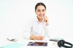 Asian working woman sits in her office, looks at tablet and writes down information, isolated on white background photo