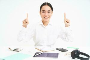 Business and profession. Portrait of asian woman in workplace, sits in office and points up, shows banner advertisement, white background photo