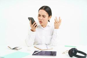 Angry lady boss, businesswoman looks frustrated, talks on mobile phone, has an argument during conversation on cellphone, sits in her office, white background photo