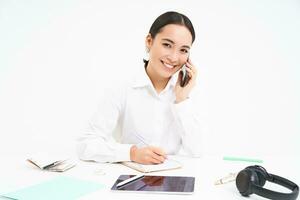 Business and company concept. Young asian woman, manager working in office, writing down information and talking with client on mobile phone, white background photo