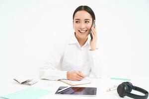 Business and company concept. Young asian woman, manager working in office, writing down information and talking with client on mobile phone, white background photo