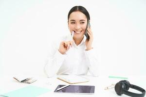 Business and company concept. Young asian woman, manager working in office, writing down information and talking with client on mobile phone, white background photo