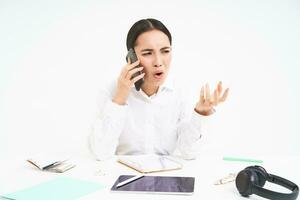 Angry lady boss, businesswoman looks frustrated, talks on mobile phone, has an argument during conversation on cellphone, sits in her office, white background photo