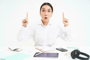 Business and profession. Portrait of asian woman in workplace, sits in office and points up, shows banner advertisement, white background photo