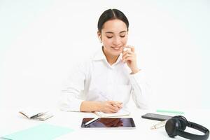 Asian working woman sits in her office, looks at tablet and writes down information, isolated on white background photo