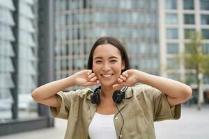 People. Portrait of smiling asian girl with headphones, dancing on street of city centre, looking happy photo