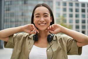 Portrait of smiling asian girl with headphones, posing in city centre, listening music photo