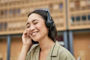 Portrait of beautiful asian woman in headphones, listening music on street of city centre, smiling happily photo