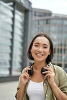 People. Portrait of smiling asian girl with headphones, dancing on street of city centre, looking happy photo