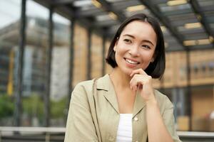 Portrait of beautiful young woman in casual clothes, smiling, posing outdoors on an empty street near glass building photo