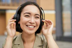 Portrait of beautiful asian woman in headphones, listening music on street of city centre, smiling happily photo