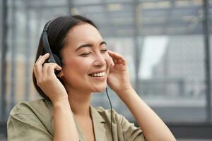 Portrait of beautiful asian woman in headphones, listening music on street of city centre, smiling happily photo