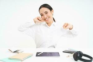 Workplace and business concept. Smiling beautiful asian woman, office worker sits in office, stretches her hands with pleased face after completed work, white background photo