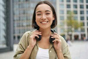 Portrait of smiling asian girl with headphones, posing in city centre, listening music photo