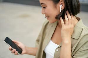cerca arriba retrato de asiático chica, mirando a móvil pantalla, escuchando música en auriculares. mujer con auriculares camina en calle foto