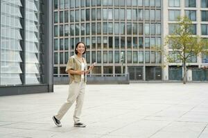 Portrait of beautiful smiling asian woman, walking on street of city centre with smartphone, looking at camera photo