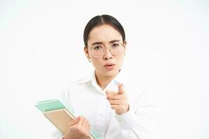 Strict asian female teacher, scolding student, pointing finger with threatening angry face, standing over white background photo