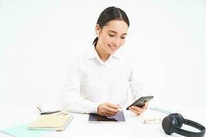 Image of businesswoman sits in office with digital tablet and headphones, looks at smartphone, checks clients messages, calling employee, white background photo