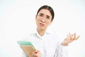 Troubled asian woman, feels complicated, looks upset and frustrated, stands with notebooks over white studio background photo