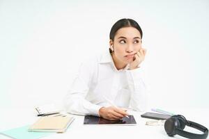 Tired and bored korean woman in office, employee sits with digital tablet, listens music in earphones and looks tired, white background photo