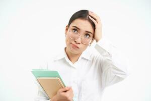 Troubled asian woman, feels complicated, looks upset and frustrated, stands with notebooks over white studio background photo