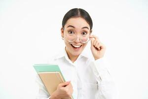 Portrait of corporate woman, asian woman in glasses, holds notebook and looks surprised, amazed by smth, stands over white background photo