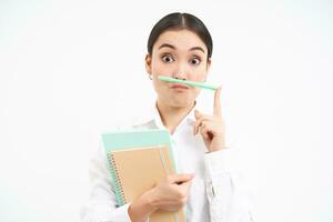 Funny korean woman with notebooks, holds pen with lips and nose and makes faces, stands over white studio background photo