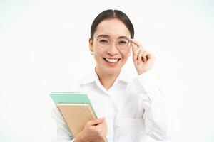 sonriente mujer profesional, asiático hembra profesor con anteojos, mirando seguro, en pie terminado blanco antecedentes foto