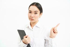 Portrait of serious asian woman with digital tablet, pointing finger right, showing banner, company advertisement, standing over white background photo