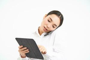 Asian working businesswoman discussing business over the phone, talking on cellphone and looking at digital tablet, standing over white background photo