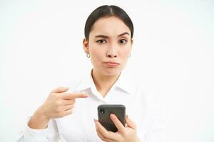 Confused businesswoman, pointing finger at smartphone and looking skeptical at camera, unsure of information online, white background photo