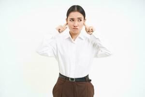 Frustrated young woman, shuts ears with fingers and grimaces from discomfort, annoyed by loud noises, white background photo