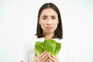 Healthy diet and raw food. Sad and unhappy asian woman, holding fresh lettuce leaves, cabbage in hands, eating vegetables, white background photo