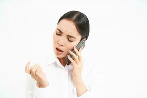 Young corporate employee, woman talks on mobile phone with lack on interest, looks at her nails while listens to person on telephone, white background photo