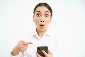 Close up portrait of happy saleswoman, successful woman with smartphone, pointing at her phone and smiling amazed, standing over white background photo