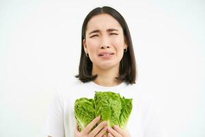 Healthy diet and raw food. Sad and unhappy asian woman, holding fresh lettuce leaves, cabbage in hands, eating vegetables, white background photo