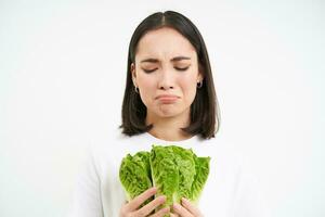 Healthy diet and raw food. Sad and unhappy asian woman, holding fresh lettuce leaves, cabbage in hands, eating vegetables, white background photo