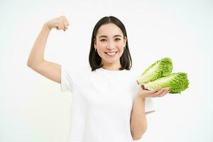 dieta y salud. sonriente asiático mujer muestra su bíceps, fuerte cuerpo y repollo en manos, come vegetales para sano nutrición, blanco antecedentes foto